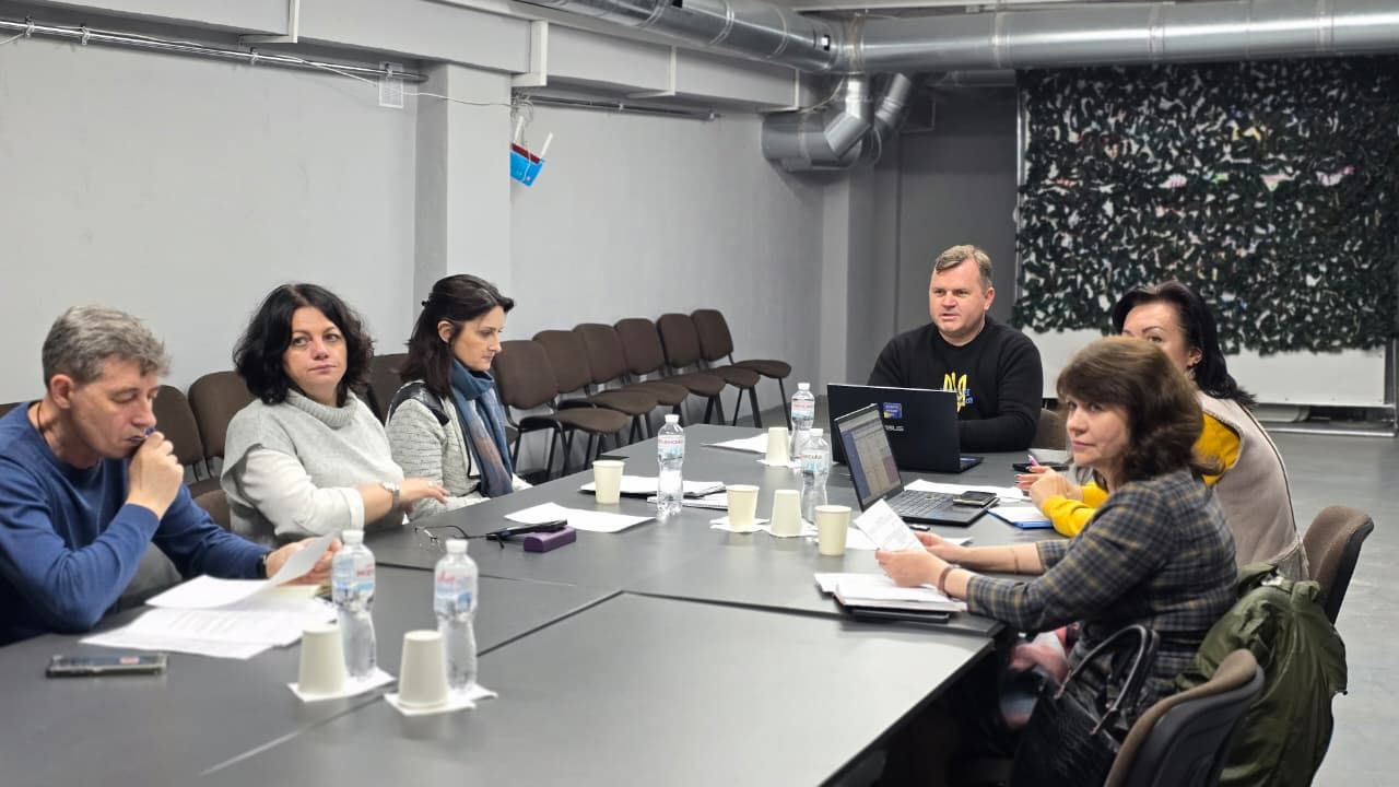 Група з шести дорослих людей (чотири жінки та двоє чоловіків) сидять за великим темним столом у сучасному конференц-залі, проводячи зустріч або обговорення. На столі видно ноутбуки, пляшки з водою та документи.