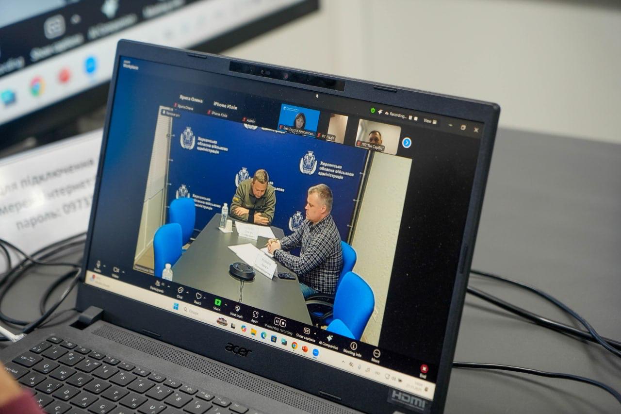 Ноутбук, на екрані якого відображається відеоконференція в Zoom. На великому вікні конференції видно двох чоловіків, які сидять за столом, а позаду них — емблема Херсонської обласної військової адміністрації.