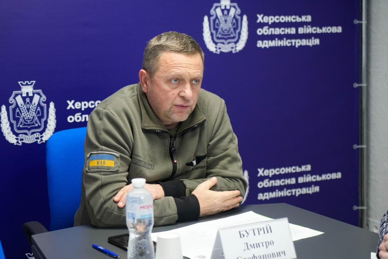 Крупний план чоловіка середнього віку в одязі військового стилю, який сидить за столом під час зустрічі. На стіні позаду нього видніється логотип і текст: Херсонська обласна військова адміністрація.