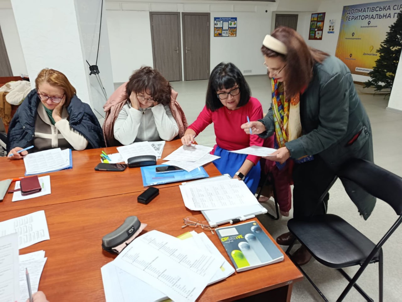 Чотири жінки беруть участь у робочому засіданні чи тренінгу за столом, вивчаючи документи. Одна з жінок (праворуч, у темно-зеленій куртці) нахилилася, щоб обговорити папери з іншими. На задньому плані видно частину банера Долматівської сільської територіальної громади та штучну ялинку.