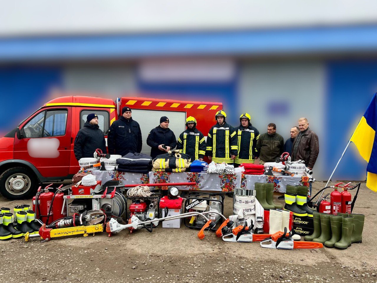 Група з восьми чоловіків, частина з яких одягнена у форму пожежників, стоїть біля червоної пожежної машини. Перед ними на столі та на землі розкладено велику кількість спеціалізованого обладнання: бензопили, мотокоси, пожежні насоси, шланги, вогнегасники та гумові чоботи. Праворуч видно український прапор.