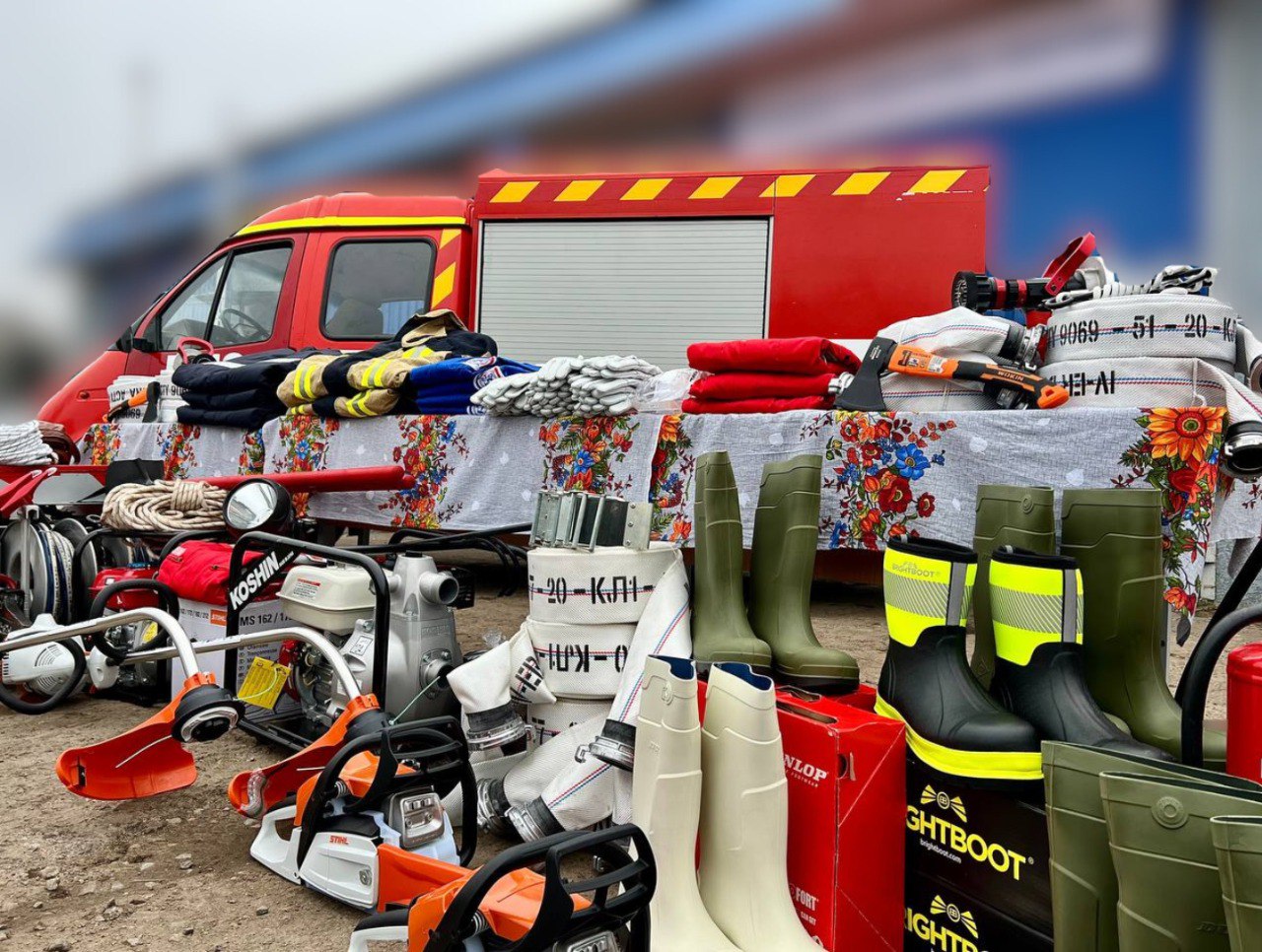 Крупний план обладнання для пожежно-рятувальних робіт, виставленого на столі та на землі перед червоною пожежною машиною. Видно мотопомпи, бензопили (зокрема Stihl), пожежні рукави, захисний одяг, рулони тканин, а також великий асортимент гумових чобіт різних кольорів.