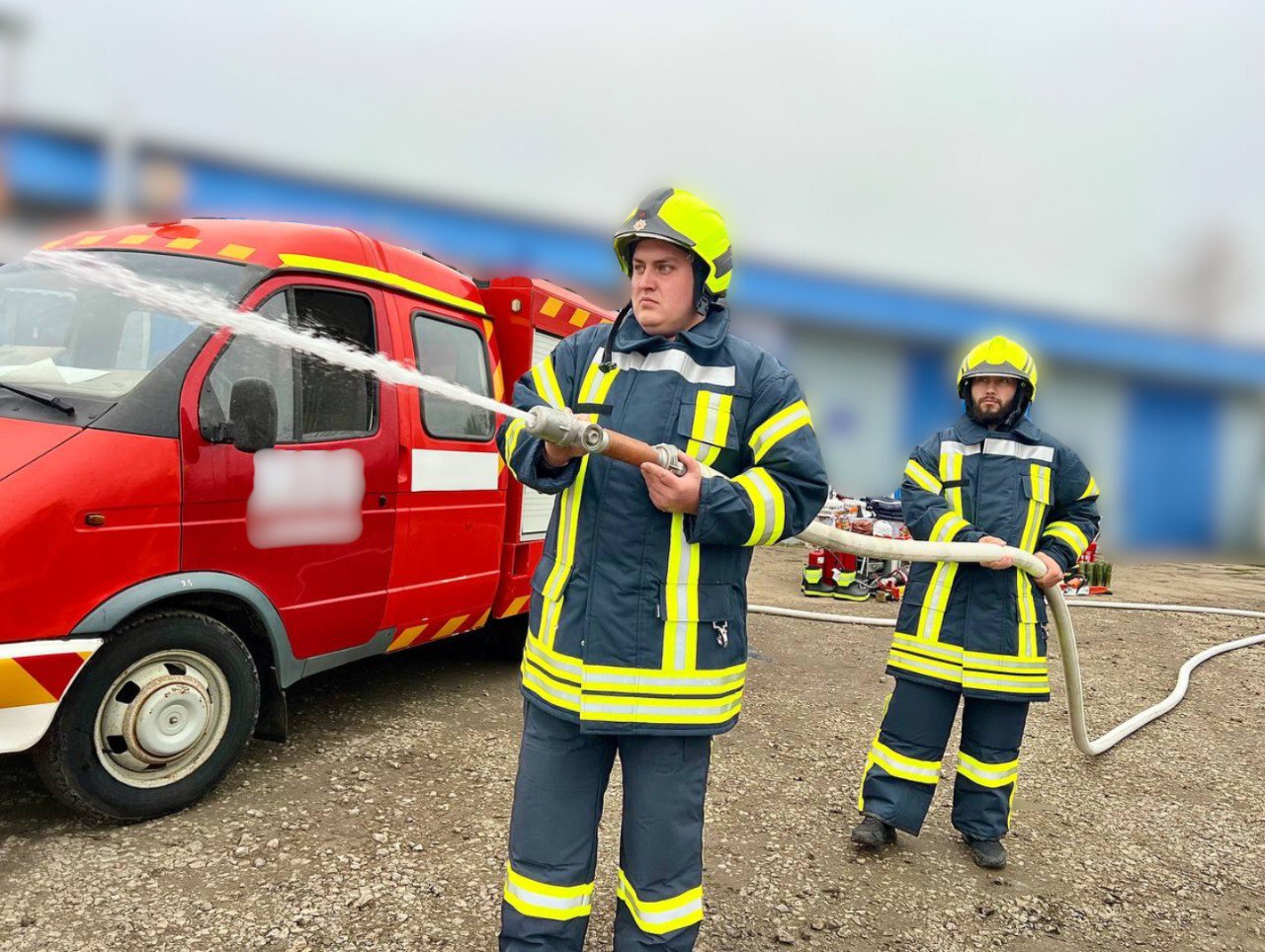 Двоє пожежників у захисних костюмах і касках стоять на вулиці біля червоної пожежної машини. Пожежник на передньому плані тримає в руках брандспойт, з якого сильно б'є струмінь води.
