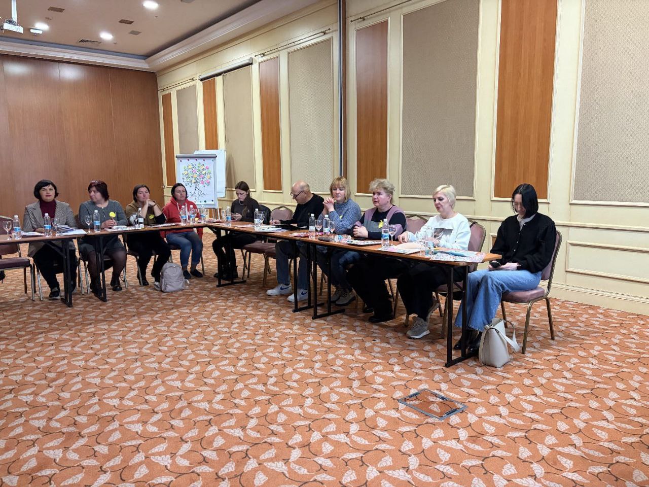 Група з десяти людей сидить за U-подібним столом у конференц-залі з килимом із візерунком, беручи участь у зустрічі або тренінгу.