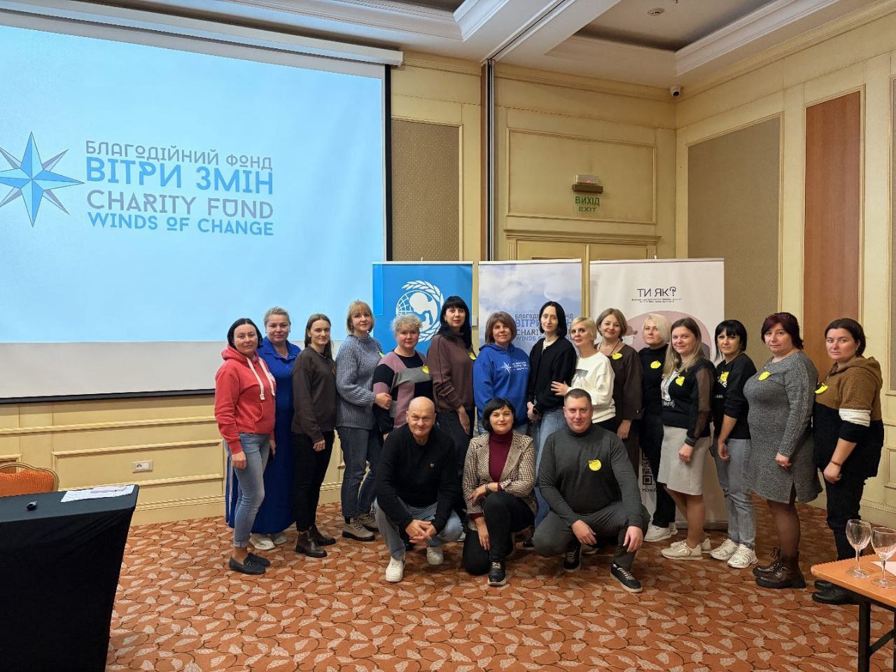 Групове фото близько 20 осіб, переважно жінок, на заході в приміщенні. Вони стоять і сидять навприсядки перед екраном із логотипом Благодійний фонд ВІТРИ ЗМІН CHARITY FUND WINDS OF CHANGE. На задньому плані також видно банери з логотипами ООН та програми ТИ ЯК? Багато учасників мають жовті наліпки.