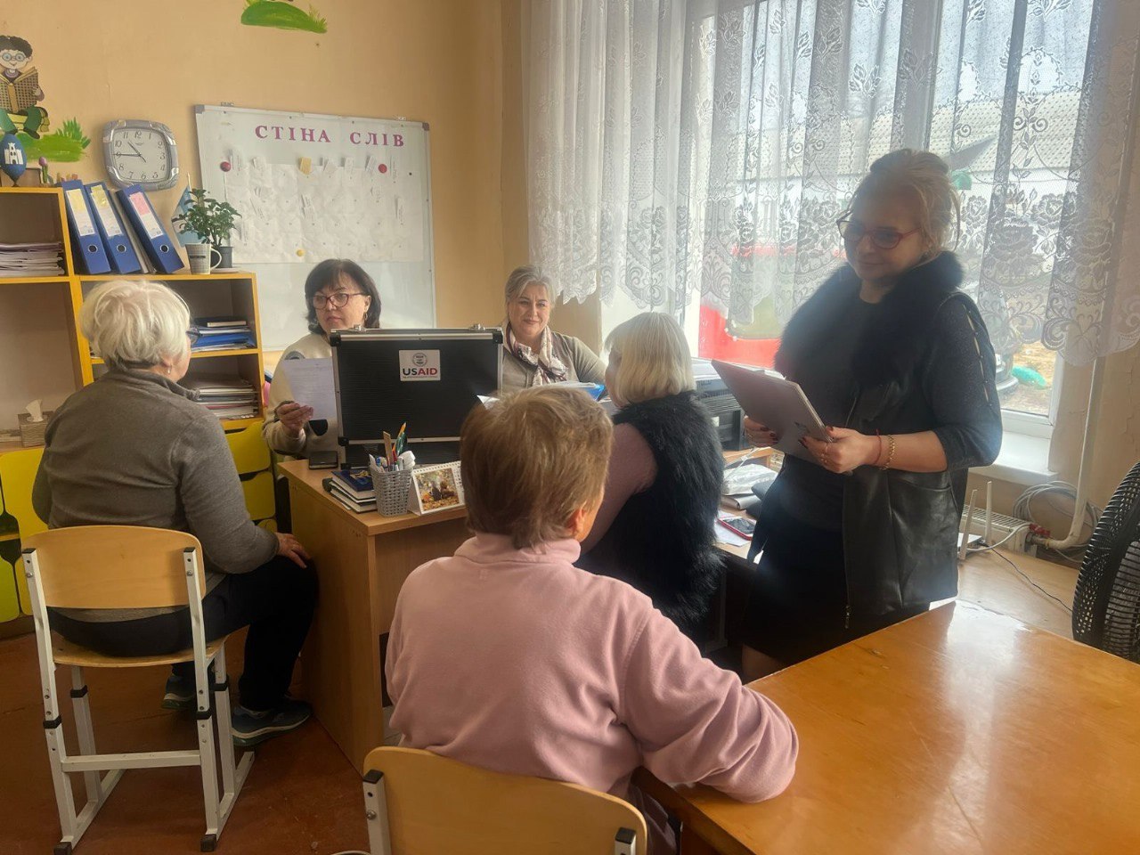 П'ять жінок перебувають у світлому офісному чи класному приміщенні. Четверо жінок зібралися навколо столу, на якому видно монітор із логотипом USAID, а п'ята жінка, що стоїть праворуч, тримає папку та щось обговорює з групою. На стіні позаду видно напис: СТІНА СЛІВ та шкільні приналежності.