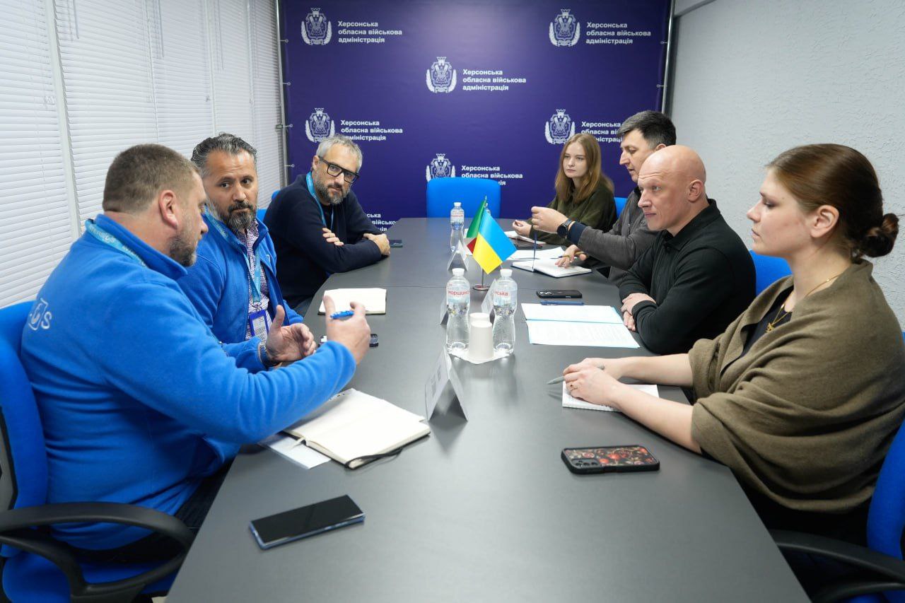 Група з семи людей, включно з двома чоловіками у синіх толстовках зліва, проводить зустріч за великим столом. На тлі — банери Херсонської обласної військової адміністрації. На столі видно пляшки води та прапорець України.