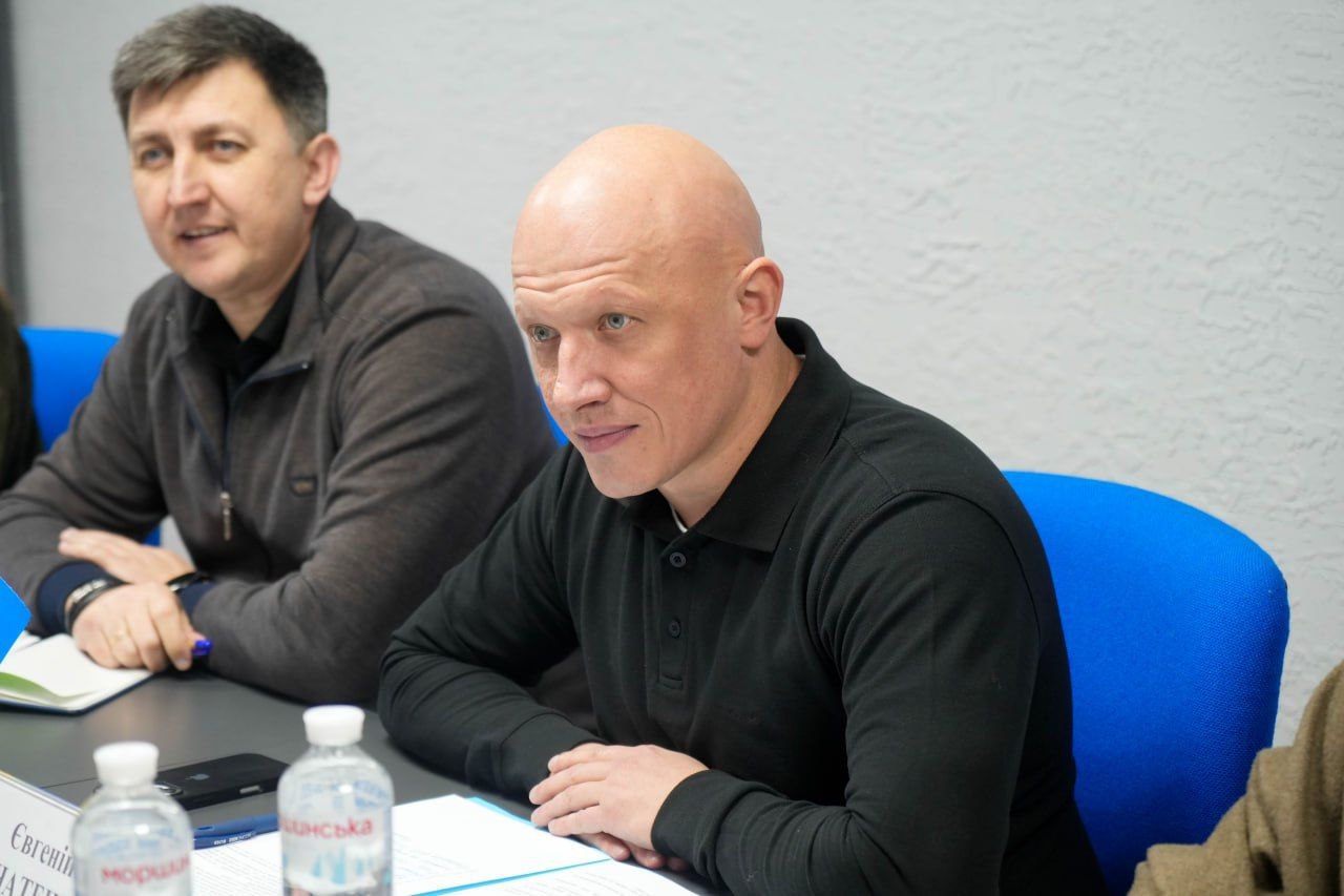 Крупний план двох чоловіків, які сидять за столом на зустрічі. На передньому плані — лисий чоловік у чорній сорочці поло, який уважно слухає, а за ним ліворуч — інший чоловік, що посміхається.