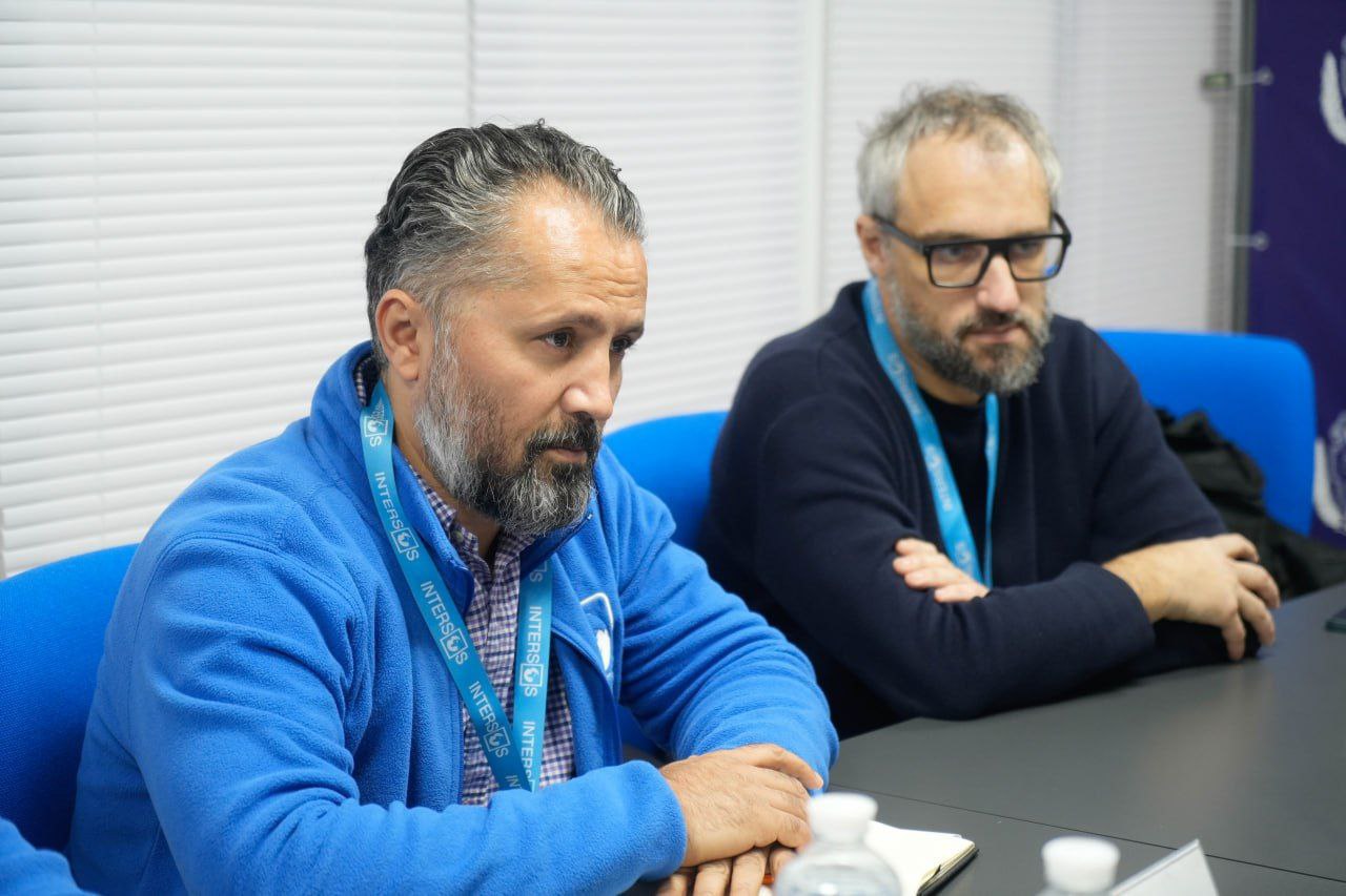 Крупний план двох чоловіків, які сидять за столом на зустрічі. На передньому плані — бородатий чоловік у синій флісовій кофті з бейджем INTERSOS, а за ним, праворуч, — інший бородатий чоловік в окулярах.