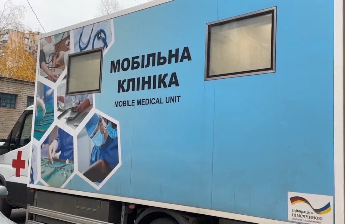 Бокова частина великого фургона або причепа, пофарбована у світло-блакитний колір, що слугує мобільною клінікою. Великими літерами написано: МОБІЛЬНА КЛІНІКА та англійською: MOBILE MEDICAL UNIT. Зліва у формі сот розташовані зображення медичних процедур. У нижньому правому куті видно напис: співпраця з Німеччиною (Deutsche Zusammenarbeit).