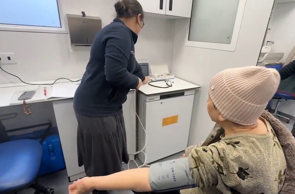 Медичний працівник (зі спини) готує обладнання для обстеження всередині мобільної клініки. На передньому плані жінка-пацієнтка в світлій шапці та камуфляжній кофті сидить на стільці. На її правій руці закріплена манжета електронного тонометра для вимірювання артеріального тиску. На задньому плані видно білі меблі та холодильник.