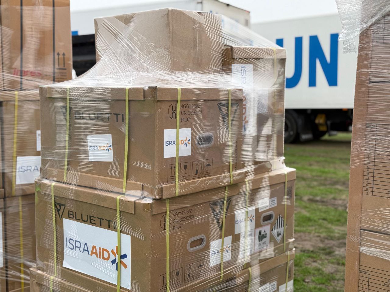 Крупний план високої палети з коричневими картонними коробками, обмотаними прозорою плівкою, які містять логотипи BLUETTI та ISRAAID. На задньому плані видно частину білої вантажівки з синіми літерами: OOH-UN.