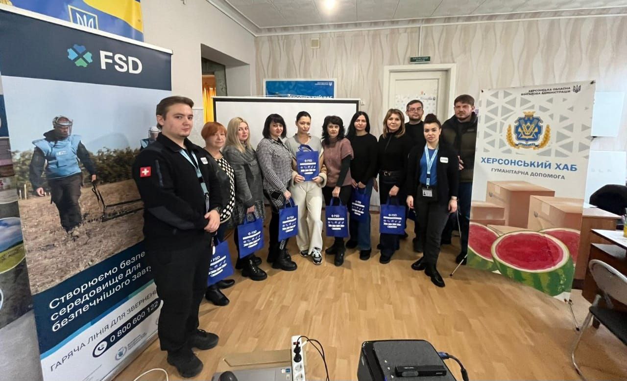 Групове фото 12 людей, які стоять у приміщенні біля банерів. Більшість із них – жінки, які тримають сині сумки. На банерах видно логотипи FSD (The Foundation for Sustainable Development), швейцарський прапор і напис: Херсонський Хаб Гуманітарна Допомога.