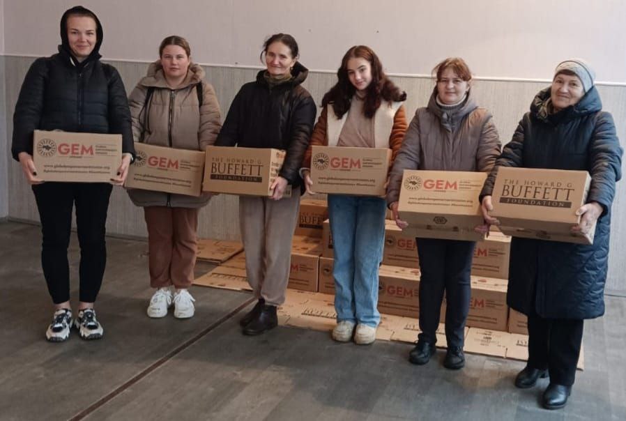 Групове фото шести жінок та дівчат, які стоять у приміщенні та тримають в руках картонні коробки гуманітарної допомоги. На коробках видно логотипи GEM (Global Empowerment Mission) та The Howard G. Buffett Foundation. Позаду них складено велику кількість таких самих коробок.