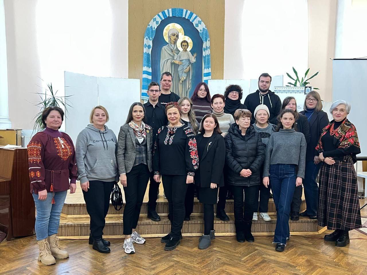 Груповий портрет приблизно 18-20 людей різного віку, які стоять на сходах у приміщенні з паркетною підлогою. На задньому плані видно велику фреску або ікону із зображенням Богородиці з Дитям. Деякі жінки одягнені у вишиванки або традиційні хустки.