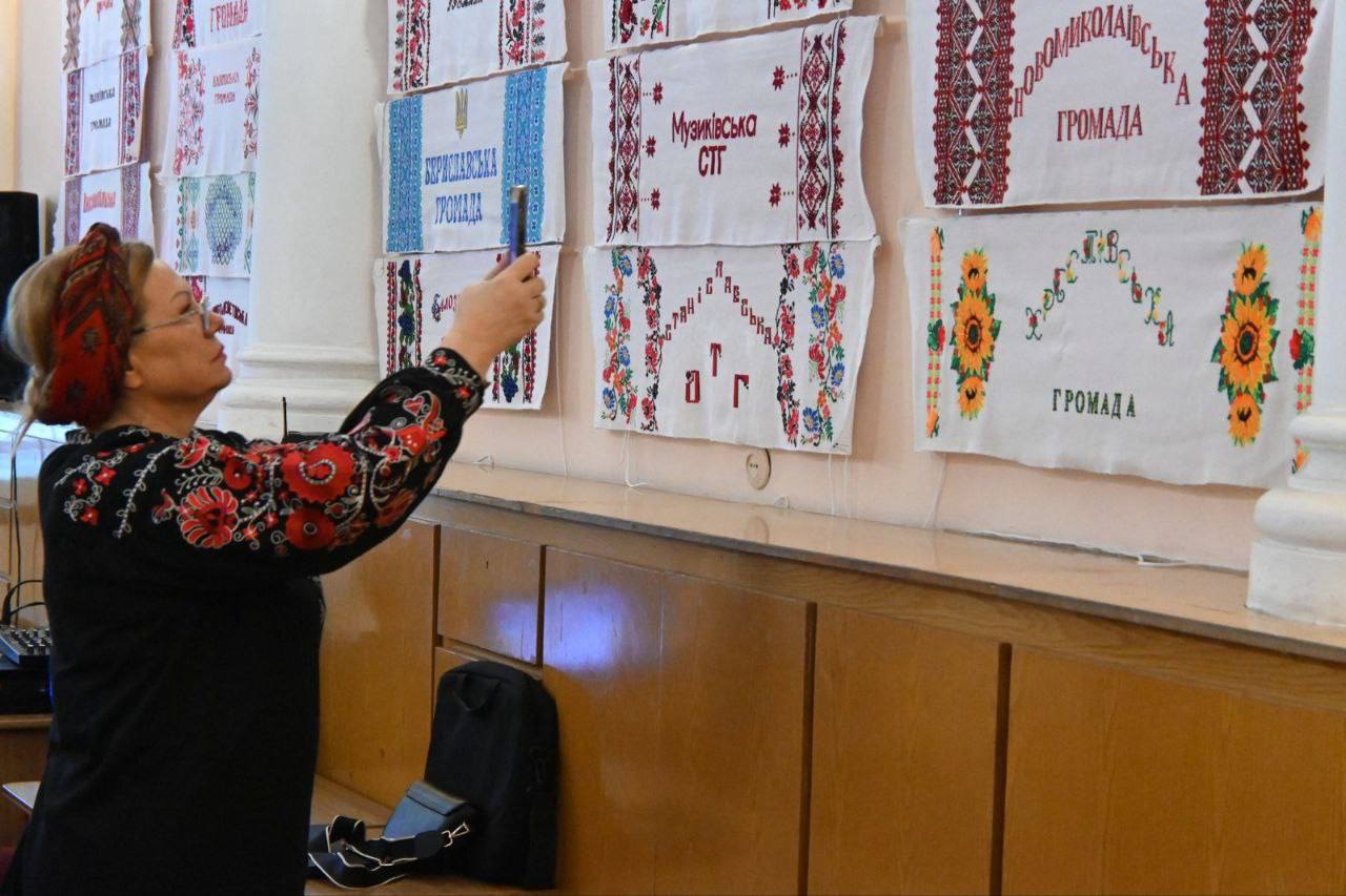 Жінка в чорній блузці з яскравою червоно-чорною вишивкою та пов'язкою на голові фотографує на мобільний телефон виставку рушників, що висять на стіні. Рушники прикрашені вишивкою та написами, що стосуються різних громад (територіальних громад), зокрема: Борозенська громада, Милівська СТГ та Великоолександрівська громада.