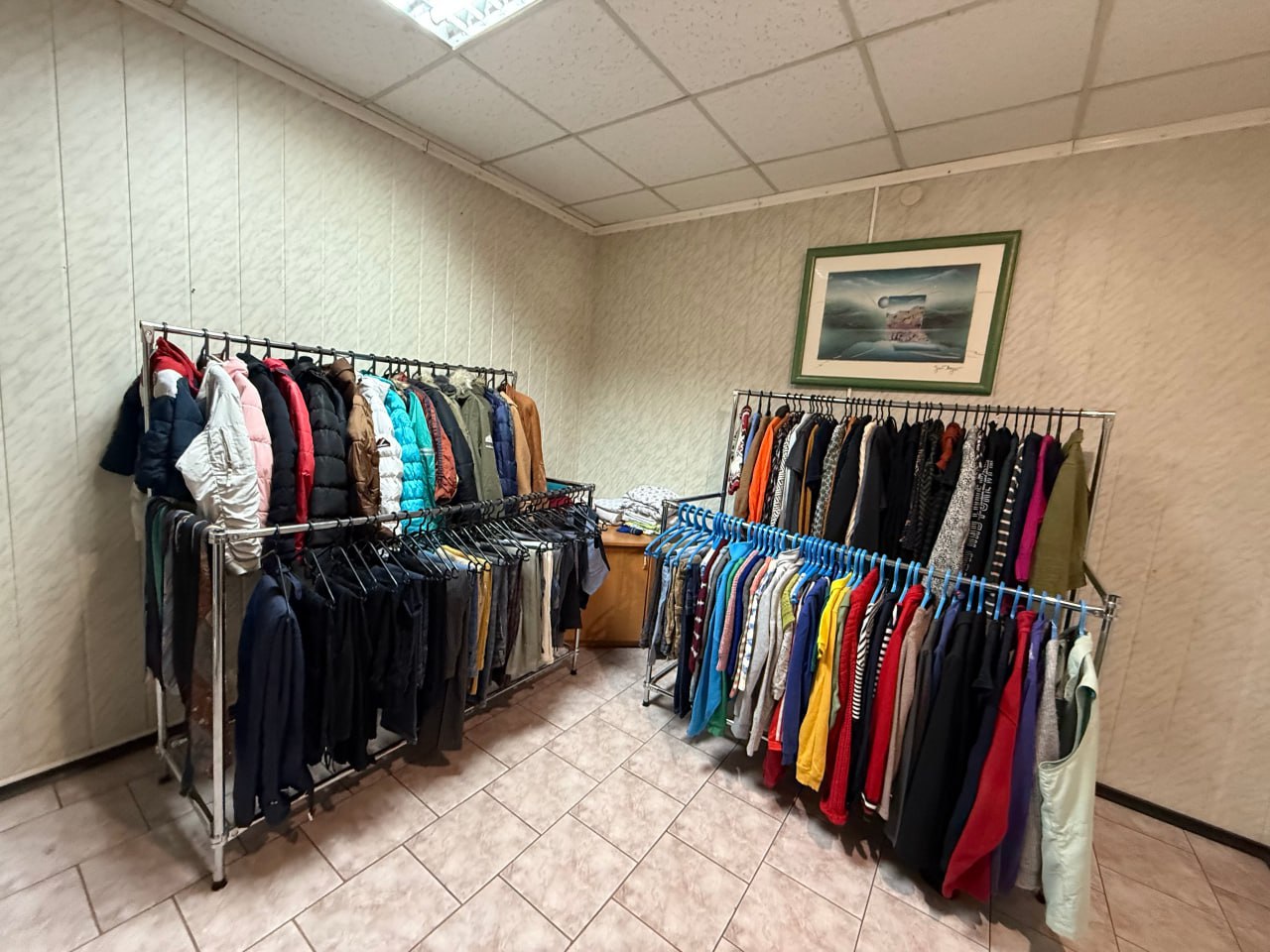 Інтер'єр невеликої кімнати з бежевими стінами та плитковою підлогою, облаштованої як склад або пункт видачі одягу. У кімнаті розміщено дві великі металеві стійки-вішалки, наповнені різноманітним одягом (куртки, светри, штани). На стіні висить невелика картина з пейзажем.