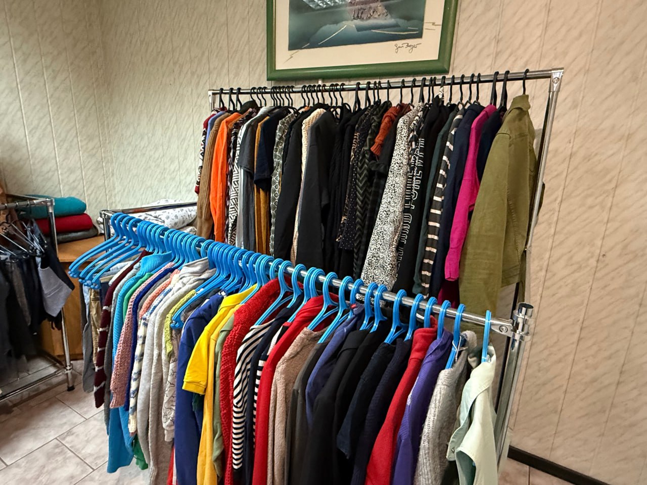 Крупний план металевої стійки-вішалки, повністю заповненої різноманітним одягом (светри, кофти, футболки) яскравих і темних кольорів на синіх і чорних плічках. Це, ймовірно, частина гуманітарної допомоги або складу одягу. На задньому плані видно частину картини на стіні.