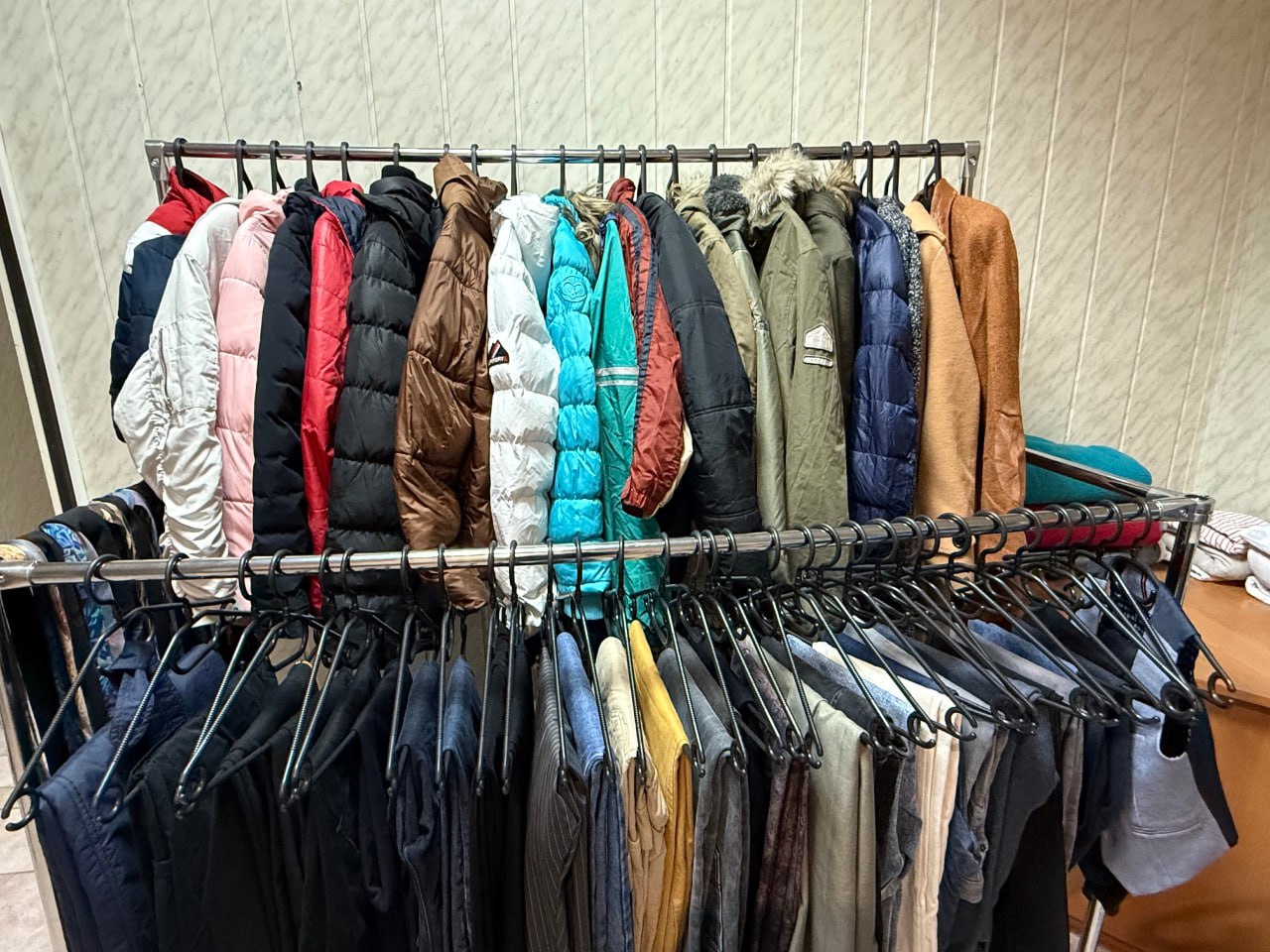 Крупний план металевої стійки-вішалки, повністю заповненої гуманітарним одягом. На верхньому ярусі висять різнокольорові зимові куртки та пуховики (червоні, білі, коричневі, бірюзові). На нижньому ярусі — штани та інший одяг темних тонів.