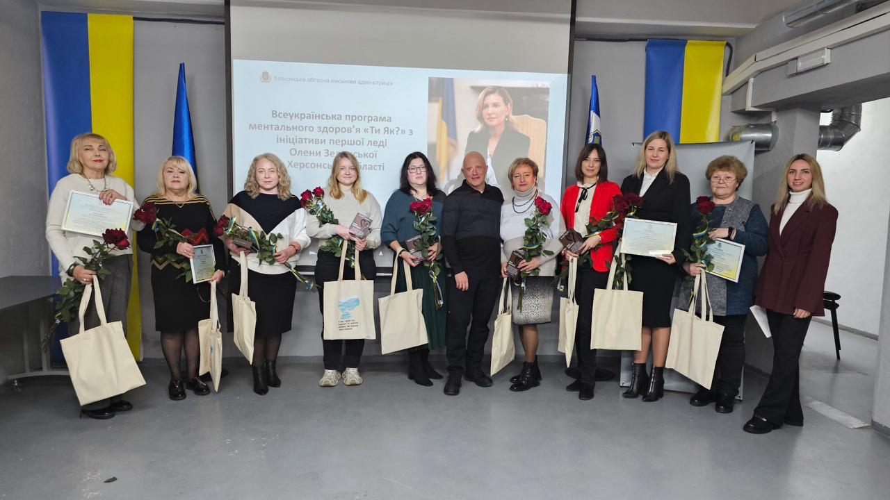 Груповий портрет одинадцяти осіб (десять жінок і один чоловік), які стоять у приміщенні на тлі екрана з написом: Всеукраїнська програма ментального здоров'я Ти як? з ініціативи першої леді Олени Зеленської. Усі зображені тримають грамоти (сертифікати), букети червоних троянд та екосумки.