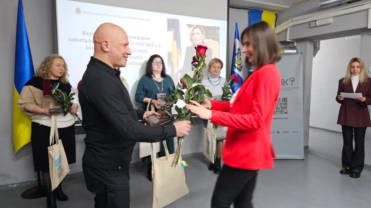 Чоловік у чорній кофті та жінка у яскраво-червоному піджаку обмінюються рукостисканням та червоною трояндою на сцені під час офіційного заходу в приміщенні. На задньому плані стоять інші жінки, а на екрані проєктора видно український текст та портрет жінки. Сцена прикрашена українськими прапорами.