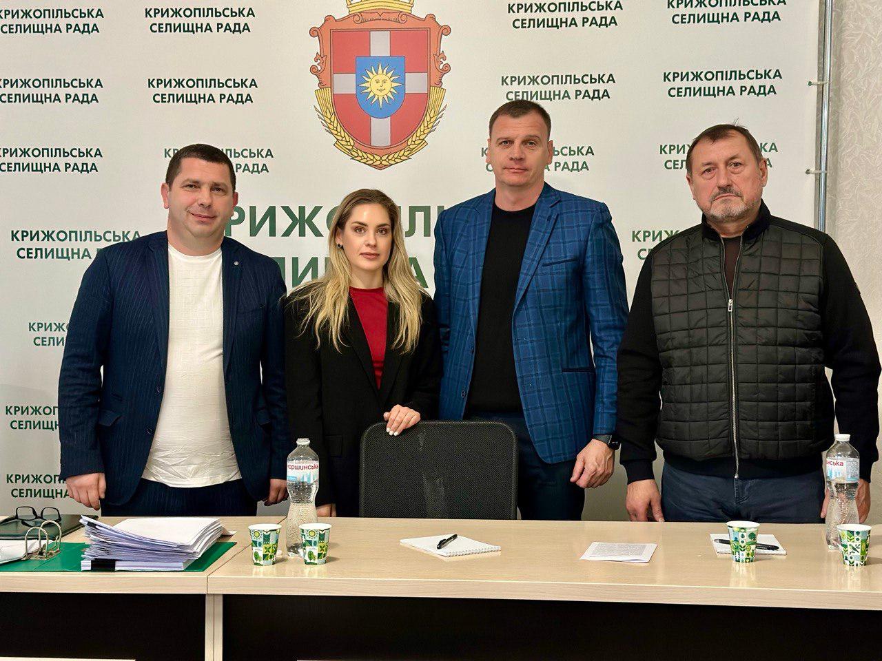 Чотири людини — двоє чоловіків у ділових костюмах, жінка в чорному піджаку з червоною блузкою та чоловік у стьобаній куртці — стоять за столом перед стіною з написом: КРИЖОПІЛЬСЬКА СЕЛИЩНА РАДА та гербом.