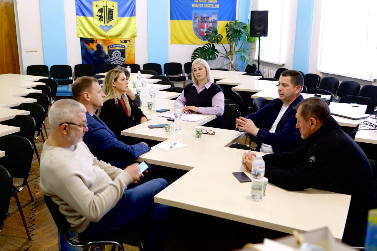 Група з шести осіб — троє чоловіків та троє жінок — сидять навколо столу в конференц-залі. На стіні за ними висять українські прапори, стяг з гербом міста та символіка Служби безпеки України.