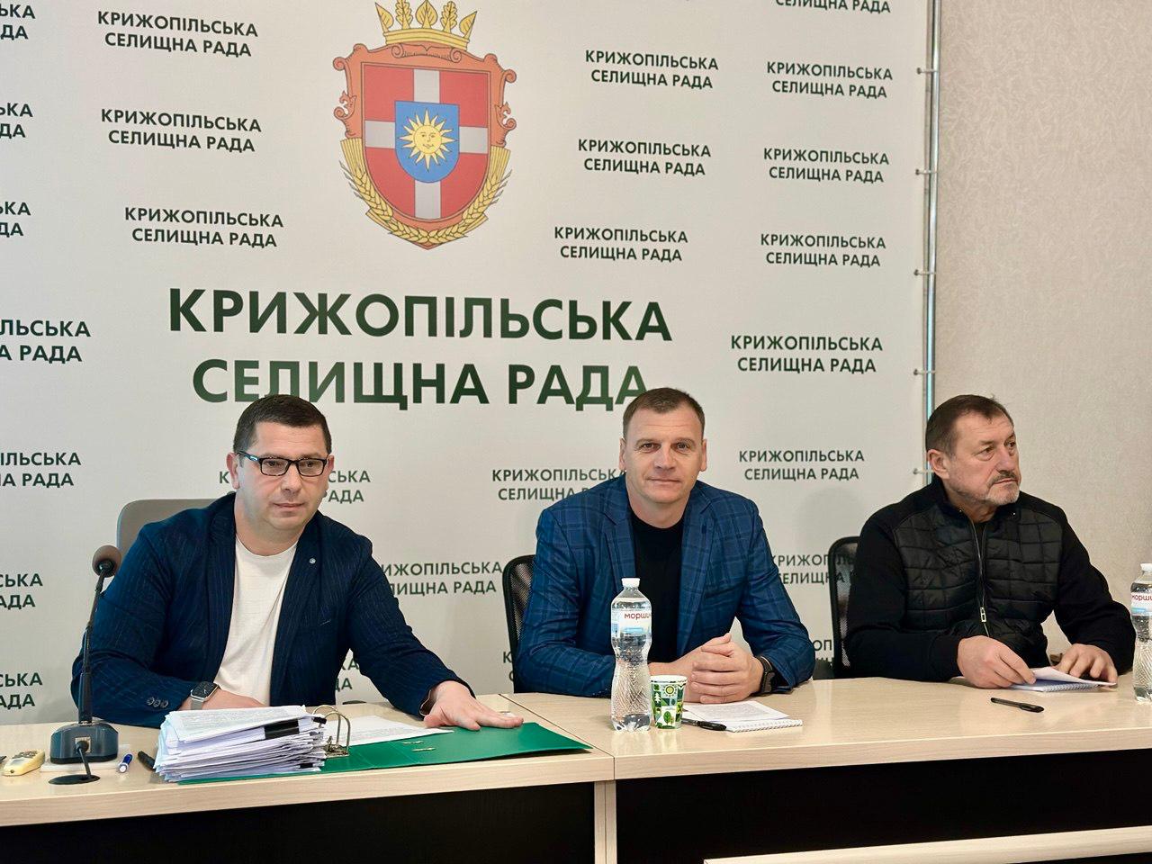 Групове фото трьох чоловіків за столом під час засідання у Крижопільській селищній раді. На фоні видно логотип селищної ради та повторювані написи: КРИЖОПІЛЬСЬКА СЕЛИЩНА РАДА.