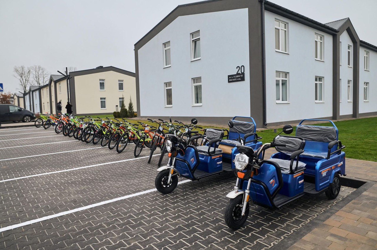Ряд яскравих двоколісних і двомісних триколісних електричних велосипедів/мопедів, більшість з яких помаранчевого, зеленого та синього кольорів, припарковані на брукованій парковці перед сучасними двоповерховими будинками. 