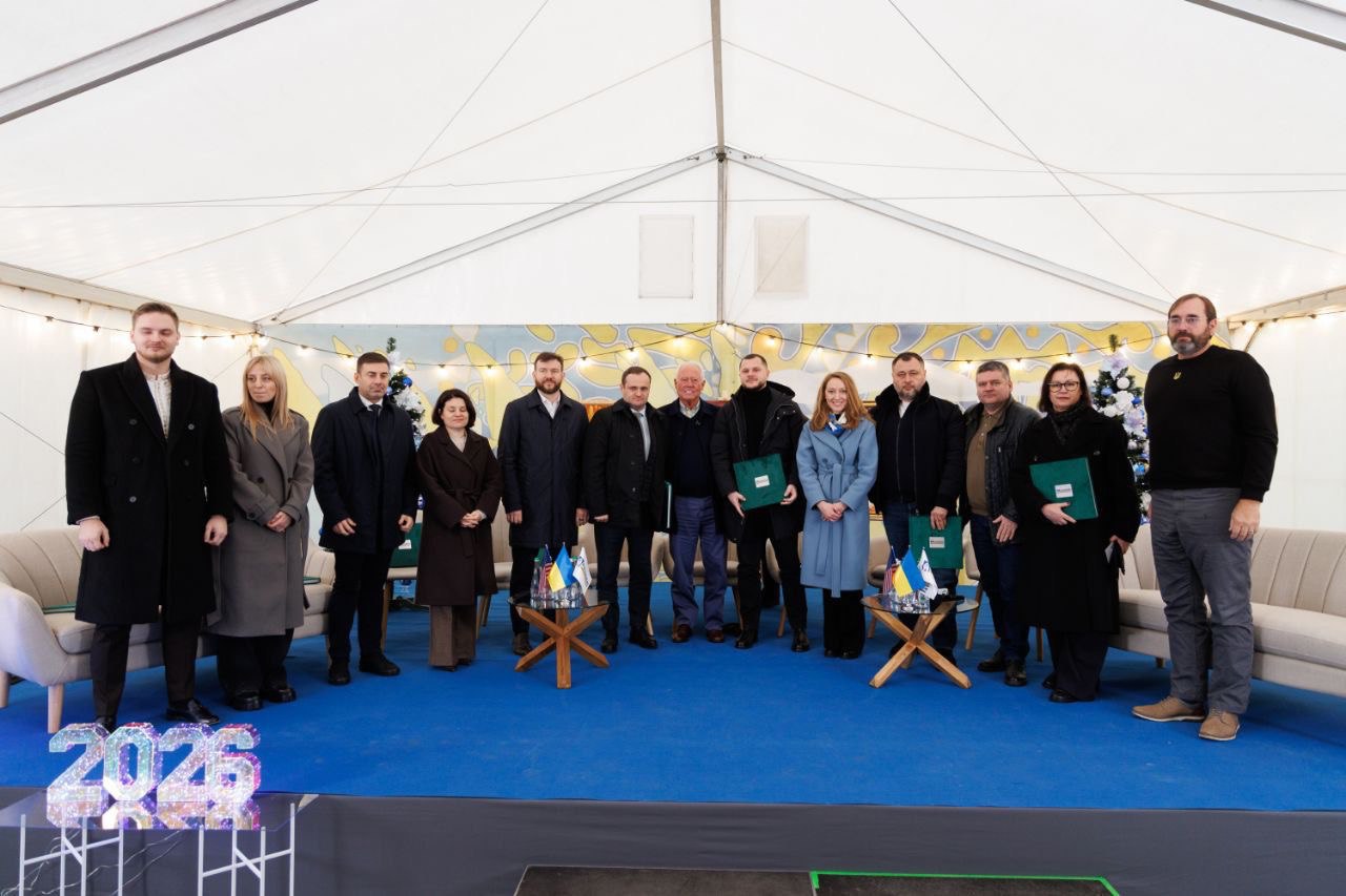Група з 13 людей, одягнених у темний одяг, стоїть під великим білим наметом. Вони розташовані в ряд, дивлячись на камеру, а двоє чоловіків у центрі тримають зелені папки. Перед ними стоять два невеликі дерев'яні столики з українськими прапорами. На передньому плані видно великі цифри 2025.
