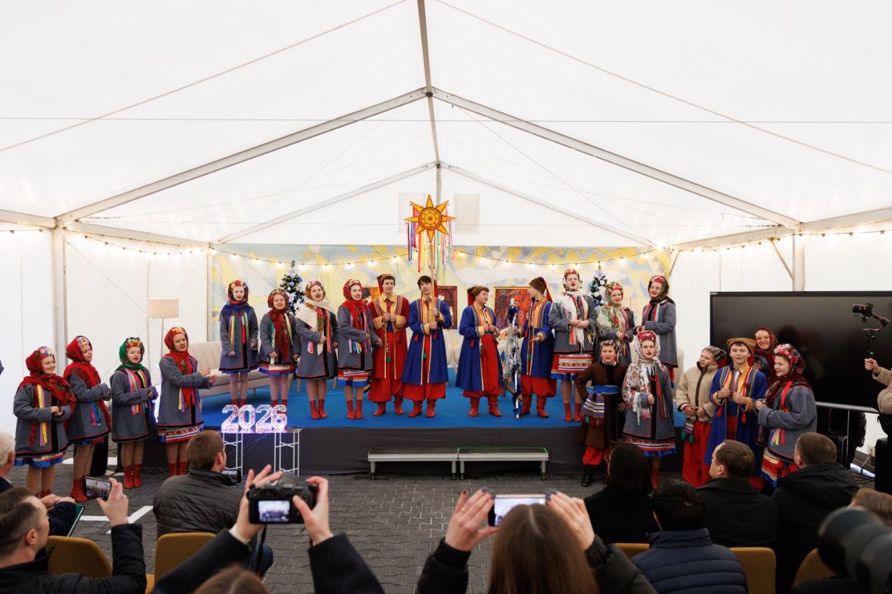 Група дітей та підлітків у традиційному українському народному вбранні — червоних, синіх та сірих пальтах і хустках — виступає на сцені під великим білим наметом. Вони стоять у два ряди, співаючи або виступаючи. Над ними висить велика різдвяна зірка. На сцені, перед виступаючими, видно великі світні цифри 2026, а внизу, на передньому плані, видно голови та руки людей, які фотографують виступ на мобільні телефони та камери.