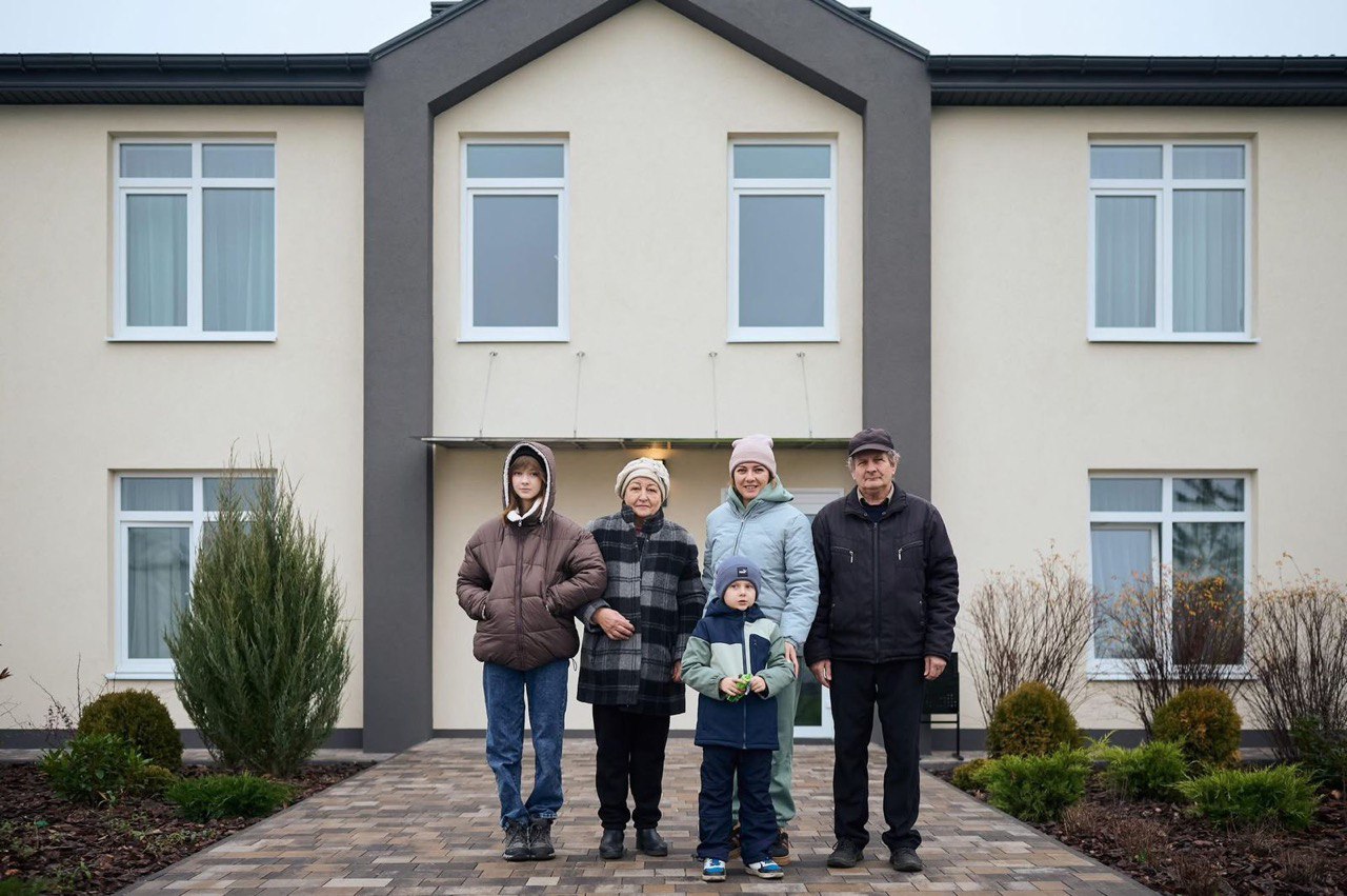 П'ять людей – троє дорослих, підліток і маленька дитина – стоять перед фасадом сучасного двоповерхового будинку, виконаного в бежевому та темно-сірому кольорах. Усі вони одягнені в зимовий одяг і дивляться прямо в камеру. Найменша дитина стоїть перед молодою жінкою у світло-блакитній куртці, а старша жінка у картатому пальті стоїть ліворуч від неї.