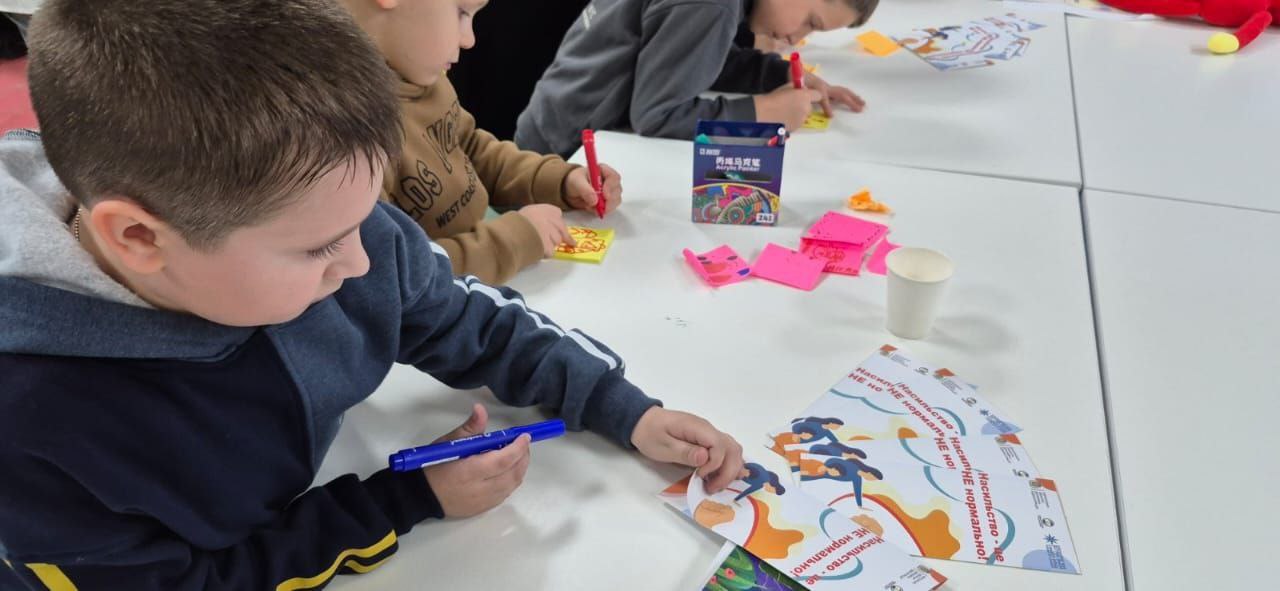 Діти сидять за білим столом, розмальовуючи маркерами великі аркуші паперу з яскравими малюнками та текстом.