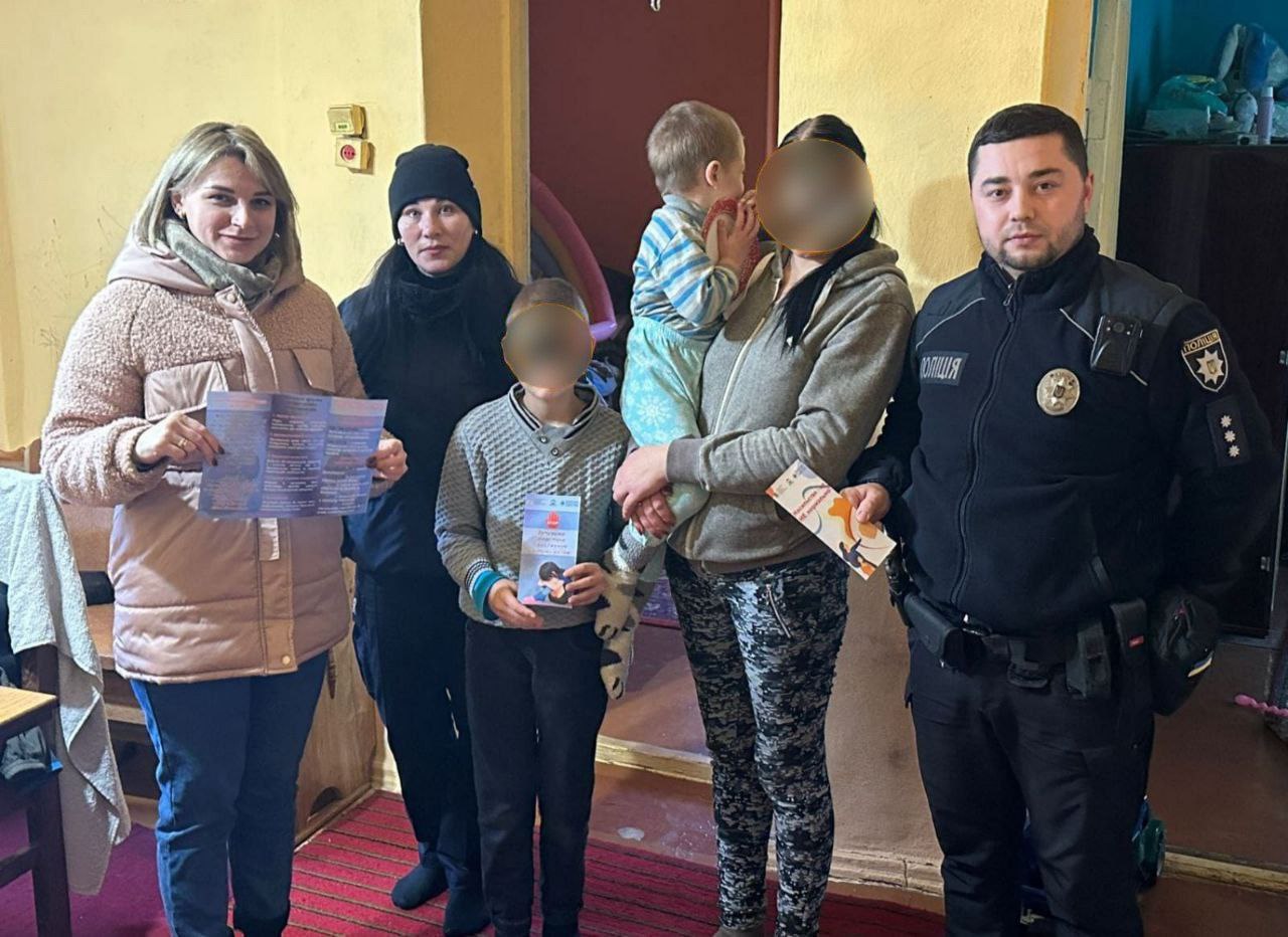 На зображенні п'ять дорослих (дві жінки та один чоловік у формі поліції праворуч) та двоє дітей. Чоловік у формі має нашивку ПОЛІЦІЯ. Дві жінки ліворуч, одна з яких тримає брошуру. Одна жінка в центрі тримає немовля, а поруч стоїть старша дитина, яка також тримає брошуру. Усі учасники стоять у дверному отворі або коридорі житлового приміщення.