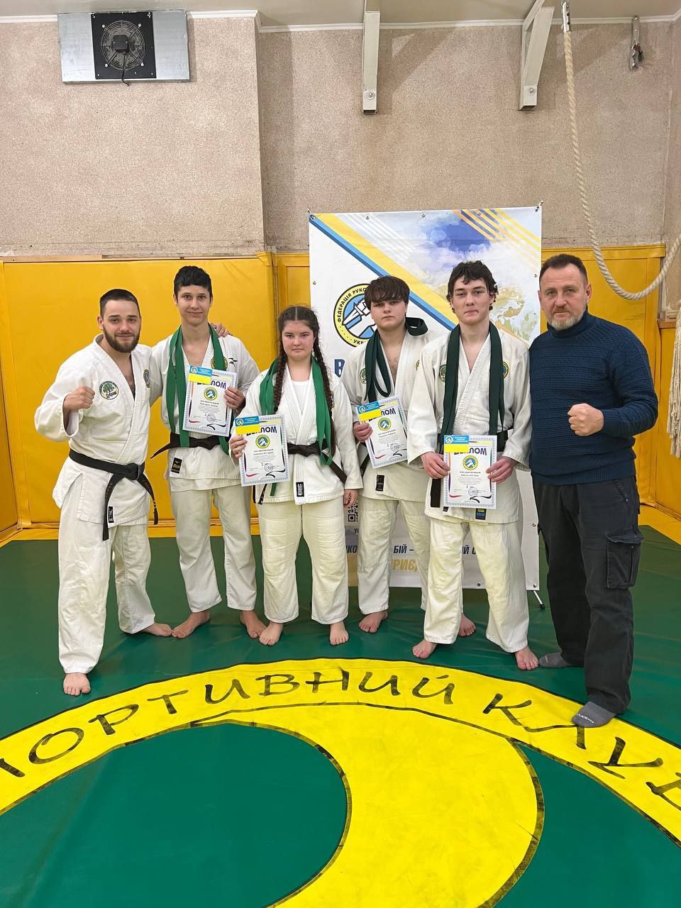 Групове фото п'яти молодих спортсменів у білих кімоно із зеленими поясами, які тримають дипломи, разом з тренером у синьому светрі, після змагань з єдиноборств.