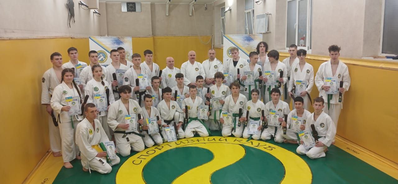 Групове фото близько 25 молодих спортсменів і двох дорослих у кімоно, більшість з яких тримають дипломи та згорнуті чорні предмети (можливо, пояси), після спортивних змагань на килимі з написом: СПОРТИВНИЙ КЛУБ.