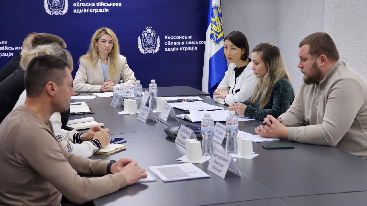 Група з восьми осіб бере участь у діловій зустрічі за великим прямокутним столом, на тлі стіни з емблемою Херсонської обласної військової адміністрації. Учасники сидять по обидва боки столу. Жінка зі світлим волоссям у світлому піджаку сидить у центрі, дивлячись прямо перед собою. Зліва від неї сидить чоловік у бежевому светрі. Чотири інші жінки та двоє чоловіків також присутні на зустрічі. На столі є пляшки з водою, паперові стаканчики та документи.