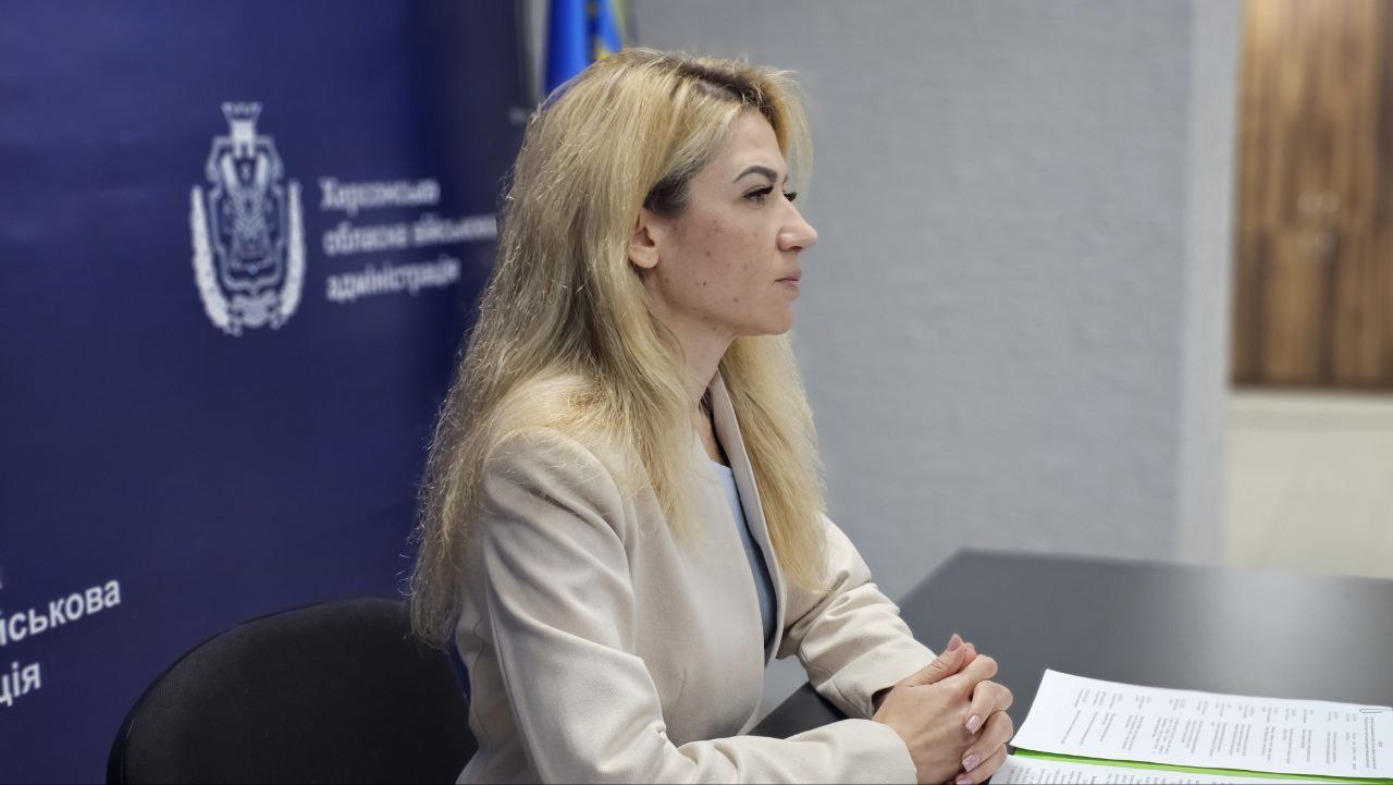 Жінка зі світлим волоссям у світлому піджаку сидить за столом у приміщенні з логотипом Херсонської обласної військової адміністрації на стіні.
