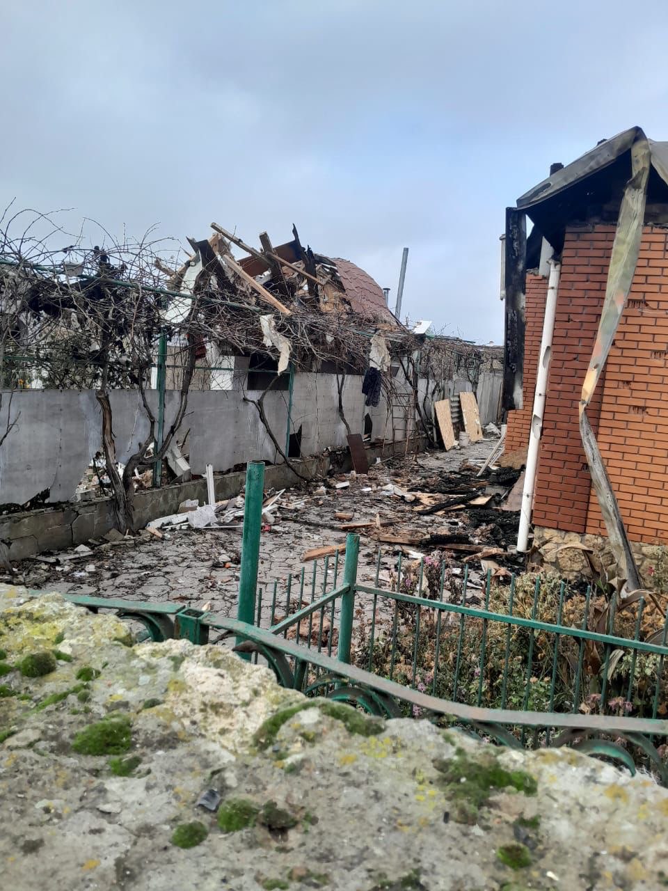 Зруйнований приватний будинок (або господарська споруда) з проваленим дахом та обгорілою цегляною стіною на подвір'ї, завалене уламками, на тлі сірого неба. Передній план займає кам'яна огорожа, вкрита мохом.