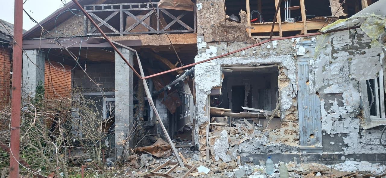 Частково зруйнований двоповерховий житловий будинок. Права сторона фасаду сильно пошкоджена: стіна розбита, видно голі балки та проріз замість вікна чи дверей, навколо якого лежать уламки штукатурки та будівельного сміття. Ліва частина будинку обгоріла, з обваленими елементами даху та обвугленими дерев'яними конструкціями на другому поверсі. Перед будинком видно сухі кущі, а на землі лежить будівельне сміття та дві пластикові пляшки.