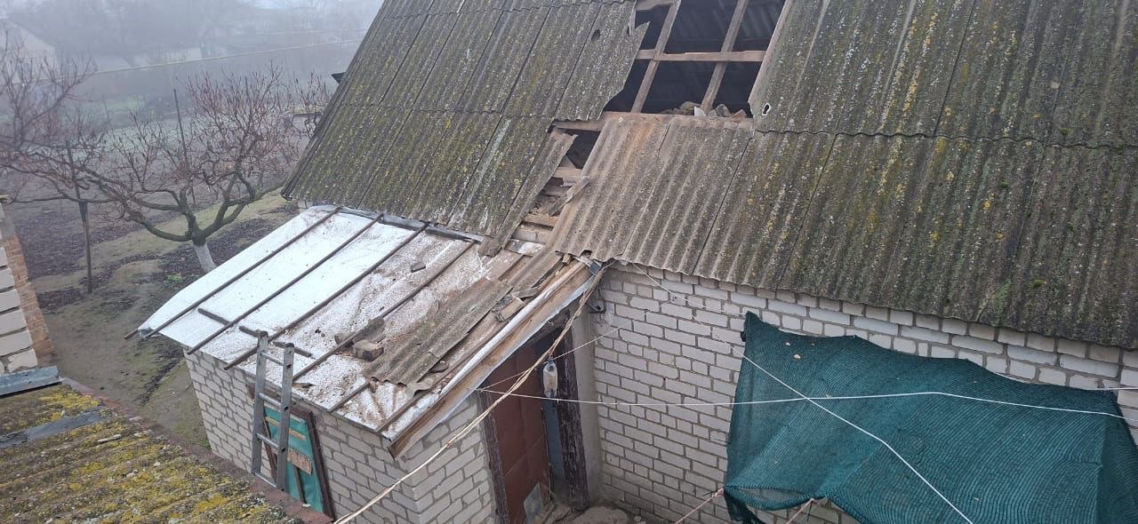 Вигляд зверху на пошкоджений дах сільського будинку. Основний дах, покритий хвилястим шифером, має значний пролом, через який видно дерев'яні балки перекриття. Нижче розташована прибудова з металевим дахом, частково присипаним снігом, притулена до стіни з білої цегли. Навколо будинку — присадибна ділянка з голими деревами, покрита туманом.