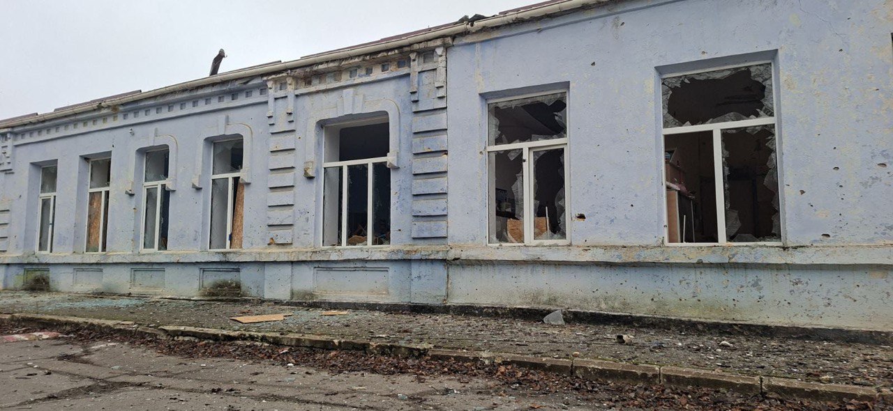 Зруйнована світло-блакитна одноповерхова будівля з численними вибитими вікнами. Деякі віконні рами повністю відсутні, інші мають розбите скло, що висить на уламках. На фасаді видно сліди пошкоджень, зокрема, від снарядів або куль. Перед будівлею знаходиться занедбана ділянка землі з розкиданим сміттям та опалим листям. Небо над будівлею похмуре.