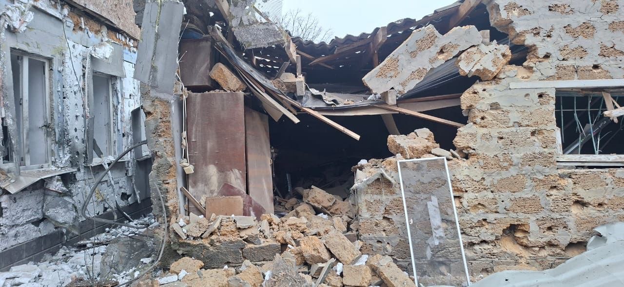 Фасад одноповерхового будинку з жовтої цегли, сильно пошкоджений вибухом або обстрілом, з великим проломом у стіні, що відкриває вид на темний інтер’єр. Частково обвалений дах із черепиці нависає над отвором. Посередині кімнати видно стару дерев’яну шафу-вітрину. Навколо пролому лежать уламки та будівельне сміття.