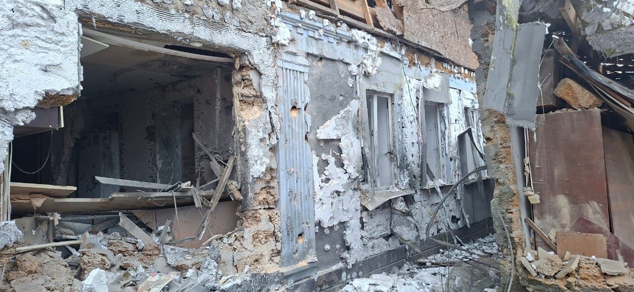 Зруйнована двоповерхова будівля зі значними пошкодженнями фасаду, вибитими вікнами та великими купами будівельного сміття та уламків на передньому плані. Видно сліди пожежі та руйнування стін і даху.