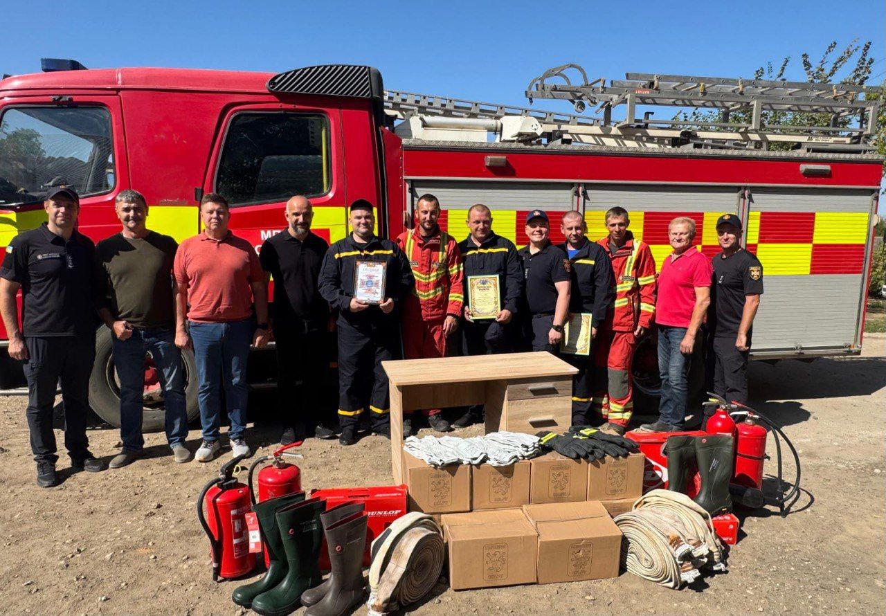 Група чоловіків, які стоять перед великою червоною пожежною машиною. Двоє чоловіків одягнені в пожежну форму, а інші – у цивільний або темний робочий одяг. Чоловіки в центрі тримають два обрамлені сертифікати або почесні грамоти. Перед ними, на землі, розташований невеликий дерев'яний стіл та розкладене пожежне обладнання: червоні вогнегасники, коробки з імовірно новою екіпіровкою, зелені чоботи Dunlop, пожежні рукави та інше приладдя. На задньому плані видно частину драбини на даху пожежної машини, яка має жовто-червону смугасту частину ззаду.