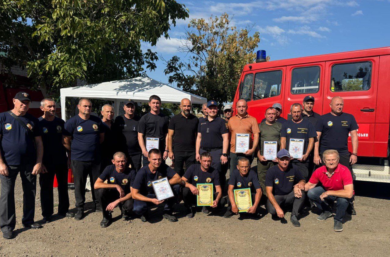 Група чоловіків, які стоять і сидять навприсядки на ґрунтовій ділянці. Чоловіки переважно одягнені у темно-сині футболки-поло з емблемами, а деякі – у цивільний одяг (чорна, оранжева, червона футболки). Чоловіки в передньому ряду (навприсядки) та деякі в задньому ряду тримають обрамлені сертифікати або почесні грамоти. На задньому плані, праворуч, видно частину червоної пожежної машини зі синім проблисковим маячком, а зліва – білий намет під зеленими деревами.