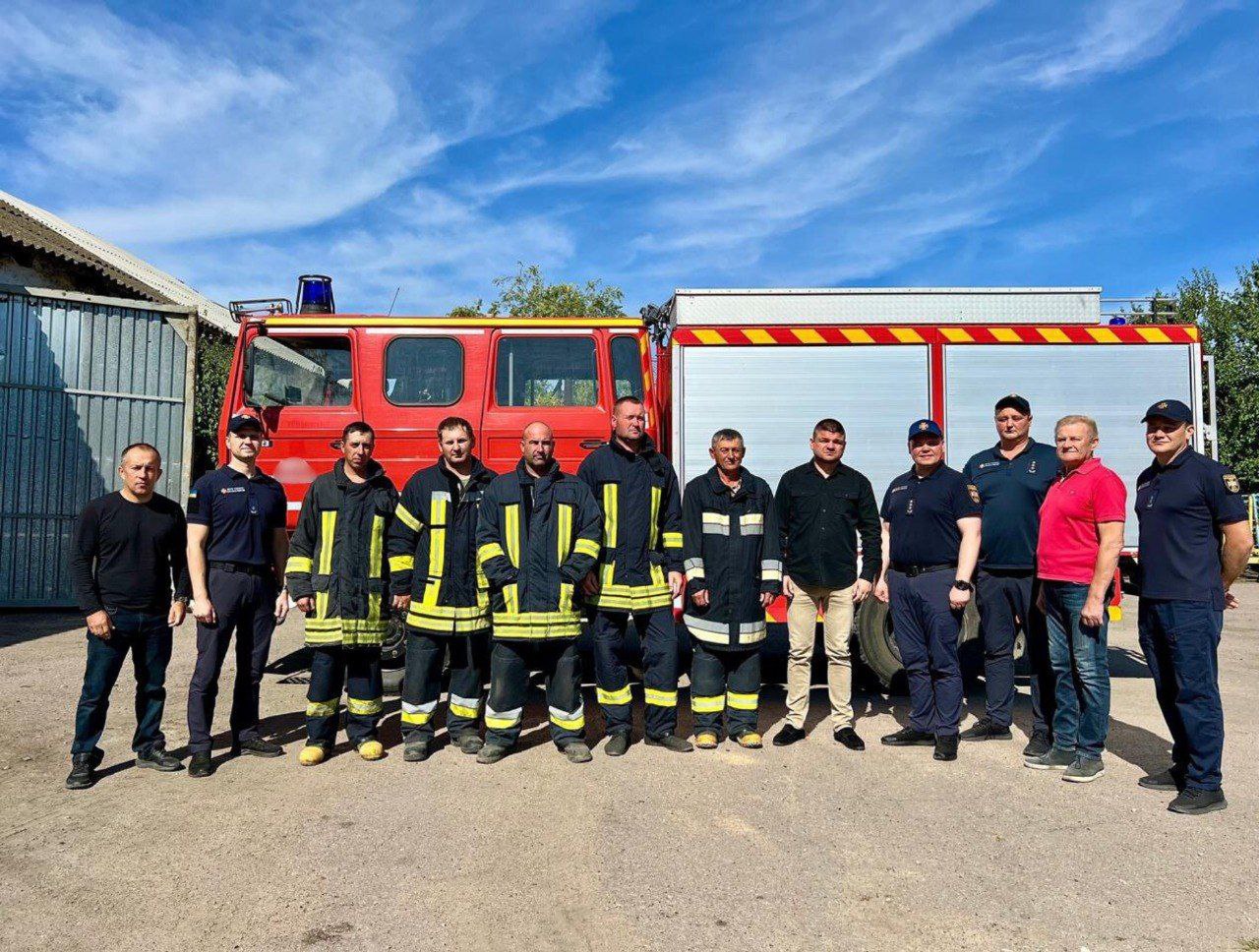 На фотографії зображено групу чоловіків, які стоять у шерензі перед червоно-білою пожежною машиною. Деякі чоловіки одягнені у повну пожежну форму з жовтими світловідбивними смугами, інші — у темно-синю або чорну робочу форму, а декілька — у повсякденний одяг. Позаду них видно частину пожежного автомобіля з резервуаром і частину металевого ангара зліва. День сонячний, небо блакитне з невеликими хмарами.