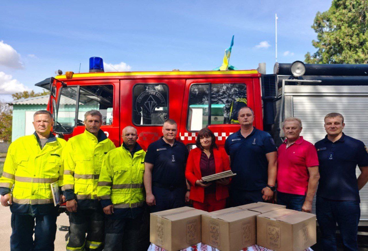 Груповий портрет восьми людей (семи чоловіків та однієї жінки), які стоять на вулиці перед червоною пожежною машиною. Жінка в червоному костюмі тримає на руках стопку нагород або документів. На столі перед ними розміщено картонні коробки. Троє чоловіків ліворуч одягнені у яскраво-жовті світловідбивні куртки.