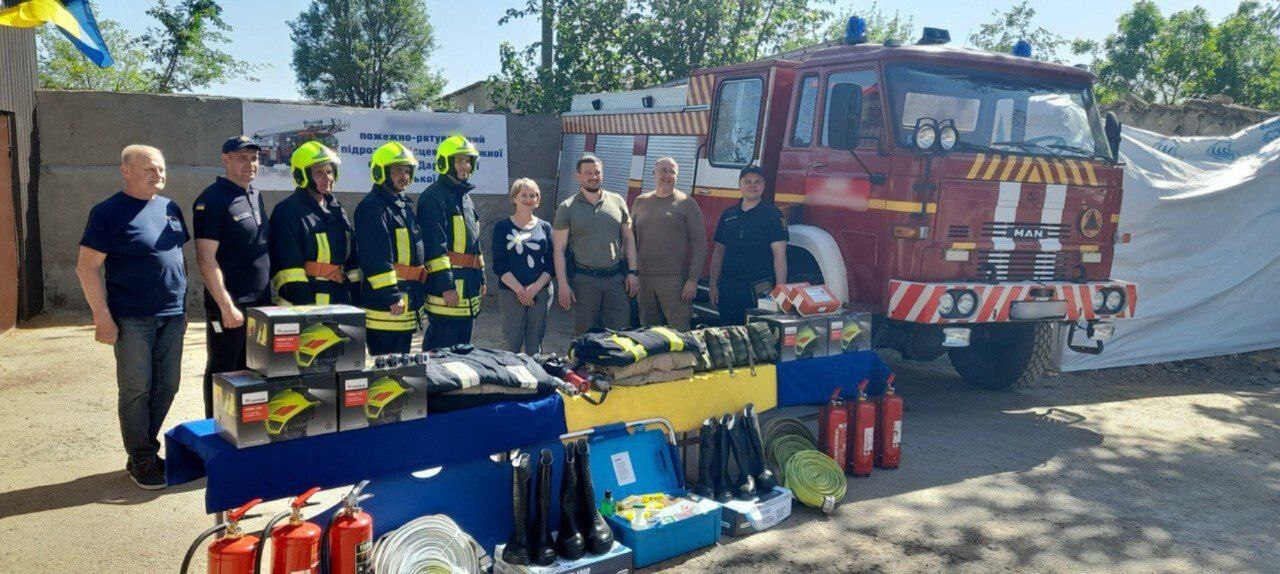 Групове фото дев'яти людей (вісім чоловіків та одна жінка), що стоять на вулиці перед пожежно-рятувальною машиною MAN. Перед ними на столі, накритому синьою скатертиною, викладено значну кількість пожежного обладнання, зокрема захисні шоломи, костюми, чоботи та вогнегасники. Частина людей одягнена в бойовий одяг пожежників, а частина — у темний та оливковий одяг. На задньому плані частково видно банер з частиною напису: пожежно-рятувальний підрозділ.