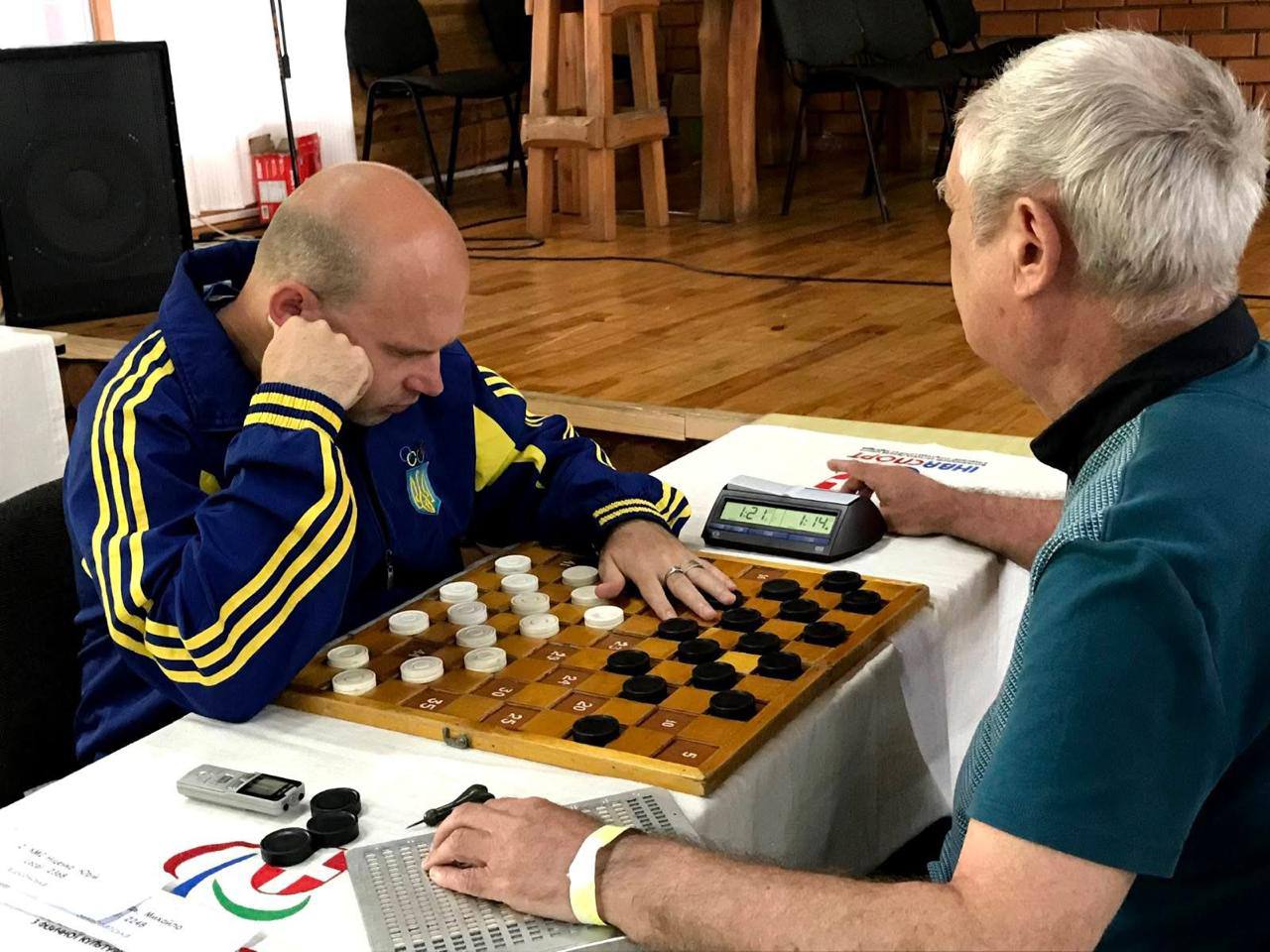 Знімок з шашкового змагання, де двоє чоловіків грають на дерев'яній дошці. Гравець ліворуч, одягнений у синю куртку з жовтими смугами (кольори українського прапора), глибоко зосереджений, підперши голову рукою. Його старший опонент у зеленуватій сорочці сидить навпроти. На столі стоїть електронний шаховий годинник (показує 1:21 і 1:14) і купа додаткових шашок. Фон — приміщення зі сценою або дерев'яними стінами.